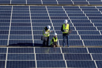 Three engineers inspect a large solar panel array together, checking equipment and performance to