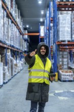 Warehouse worker in warm clothing and safety vest holding a barcode scanner and giving thumbs up