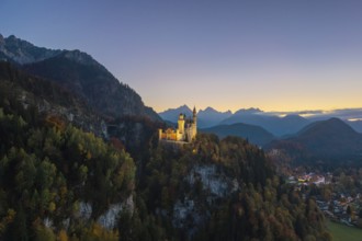 Illuminated Neuschwanstein Castle on a hill with mountain panorama under sunset-drenched sky,