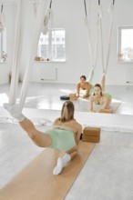 A woman is engaged in a leg stretching fitness routine using a hammock in a well-lit studio
