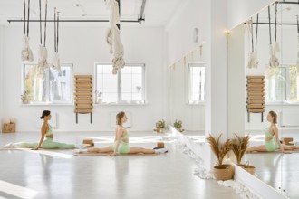 In a spacious yoga studio filled with natural light, two women perform stretches on mats. The room