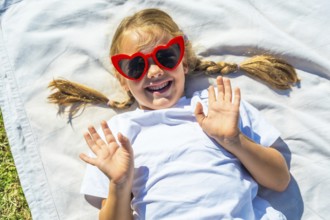 Little girl with braided blonde pigtails smiling and waving from a white blanket in the park,