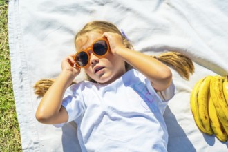 Young girl lying on a white blanket in green grass, adjusting orange sunglasses and smiling while