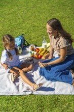 Mother and daughter share food and laughter on a picnic blanket in green grass, enjoying sunny park