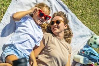Mother and daughter lying on a picnic blanket in green grass, wearing sunglasses and smiling