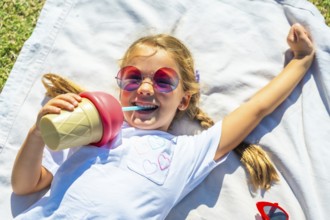 Young girl lying on a blanket in the grass on a sunny day, laughing while drinking from an ice