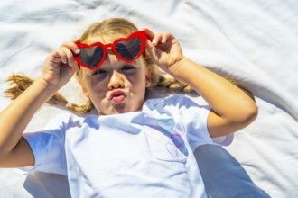 Young blonde girl lying on a white blanket making a fun kiss face while wearing red heart shaped