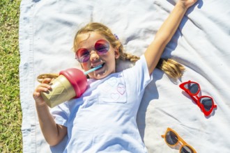 Young girl lying on a white blanket in green grass, smiling while drinking from an ice cream shaped