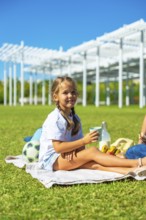 Little girl smiling, holding a cup of milk, and relaxing on a blanket during a family picnic in a