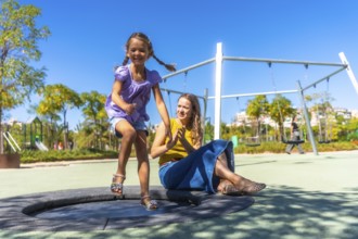 Daughter jumps on a round trampoline at a sunny park playground while mother claps nearby, sharing