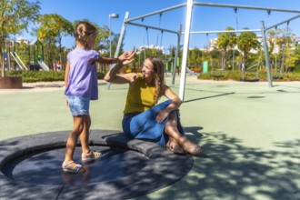 Mother and daughter high fiving on playground equipment in a sunny park, sharing a joyful bonding