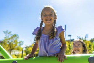 Young girl with pigtails smiling at the camera while her mother watches from playground equipment