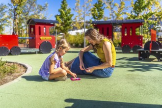 Mother and daughter bonding outdoors, sitting on the soft playground surface and concentrating on