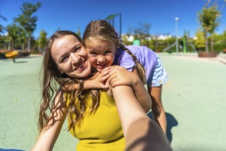 Young mother and daughter smiling at the camera while hugging on a sunny park playground, enjoying