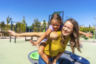 Happy mother giving her laughing daughter a piggyback ride on a sunny day in a modern public