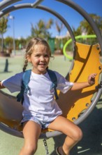 Happy little girl with a backpack smiles broadly while sitting on a swing in a vibrant outdoor