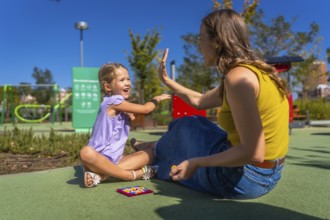 Mother and daughter sitting on playground ground, smiling and exchanging a joyful high five while
