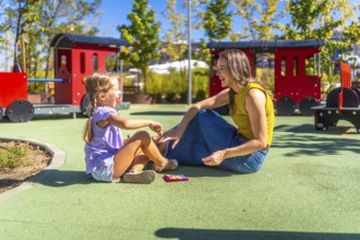 Mother and daughter sitting on soft flooring at a public playground, laughing happily during a