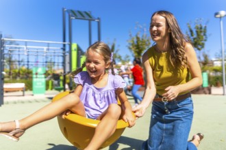 Mother and young daughter sharing laughter as mom pushes her on a bright yellow spinning cup at a