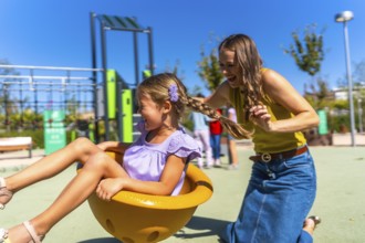Mother and daughter laughing on a sunny playground spinner, sharing carefree bonding and joyful