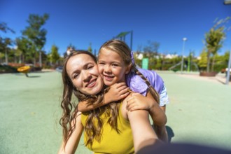 Mother and daughter smiling and embracing as they take a sunny playground selfie, enjoying a joyful