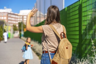 Mother waving goodbye, watching her young daughter with a backpack walk independently towards
