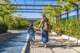 Mother and daughter, wearing backpacks, walking along a sunny pathway lined with green trees and