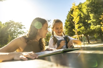 Mother and daughter smiling and laughing while reading a book together at an outdoor table in a