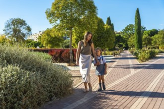 Mother and daughter walking along a path in a park, holding hands and going to school on a sunny
