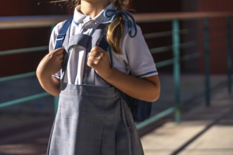 Young student in school uniform stands outdoors on a sunny day holding a backpack, ready and
