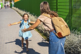 Child daughter with backpack runs across schoolyard to joyfully hug her mother at pickup,