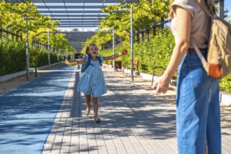 Excited schoolgirl with backpack runs toward her mother in a sunny park, holding a banana after