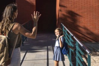 Mother waving goodbye to her elementary school daughter holding a backpack and happily walking