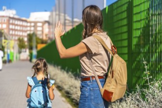 Mother waving with her hand to a small daughter carrying a backpack and walking away towards