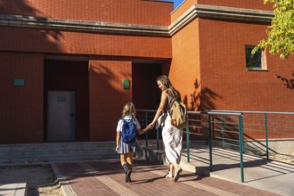 Mother holding hands with her daughter, both walking together towards a brick school building on a