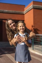 Mother and daughter smiling while standing together on a sunny day at school with the elementary