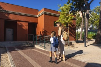 Mother and daughter smiling as they walk toward a red brick school building with backpacks,