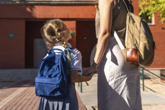 Mother and daughter walking together holding hands, wearing backpacks and school uniforms, while