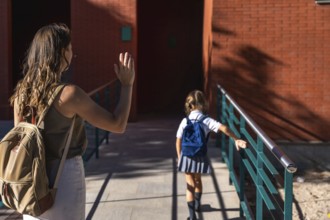 Mother waving goodbye to her elementary age daughter wearing a school uniform and backpack, walking