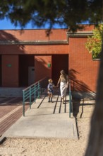 Mother and daughter holding hands and walking together, approaching a school building on a sunny