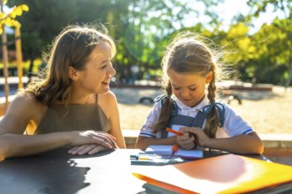 Mother and daughter sitting at a table outdoors in a park, engaging in a school activity and