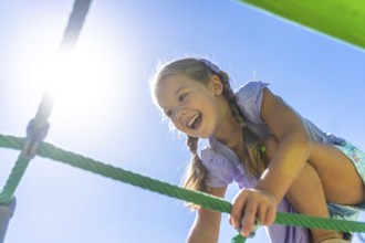 Happy young girl laughing with joy while actively climbing a green rope net structure outside on a