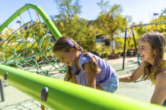 Playful daughter laughing as she climbs a green playground structure while mother smiles and