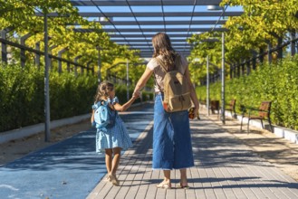 Mother and daughter holding hands and walking together along a paved path, carrying backpacks and