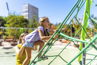 Mother assisting her happy daughter climbing a green rope jungle gym on a sunny day, experiencing