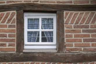 A half-timbered house window with wooden beams and blue checkered curtains, surrounded by red