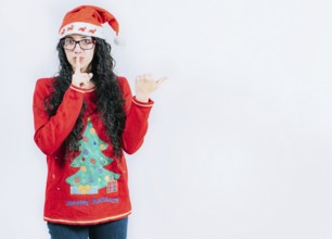 Girl in Christmas sweater and glasses pointing to the side, isolated. Young woman in Christmas hat