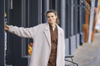 A fashionable woman is posing near a cafe entrance in a modern urban area. She wears a cozy outfit