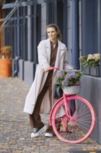 A woman stands with a pink bicycle featuring a basket filled with flowers on a city street. She is
