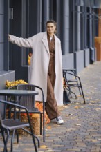 A woman stands near a sidewalk cafe, wearing a trendy outfit with a long coat. The surrounding area
